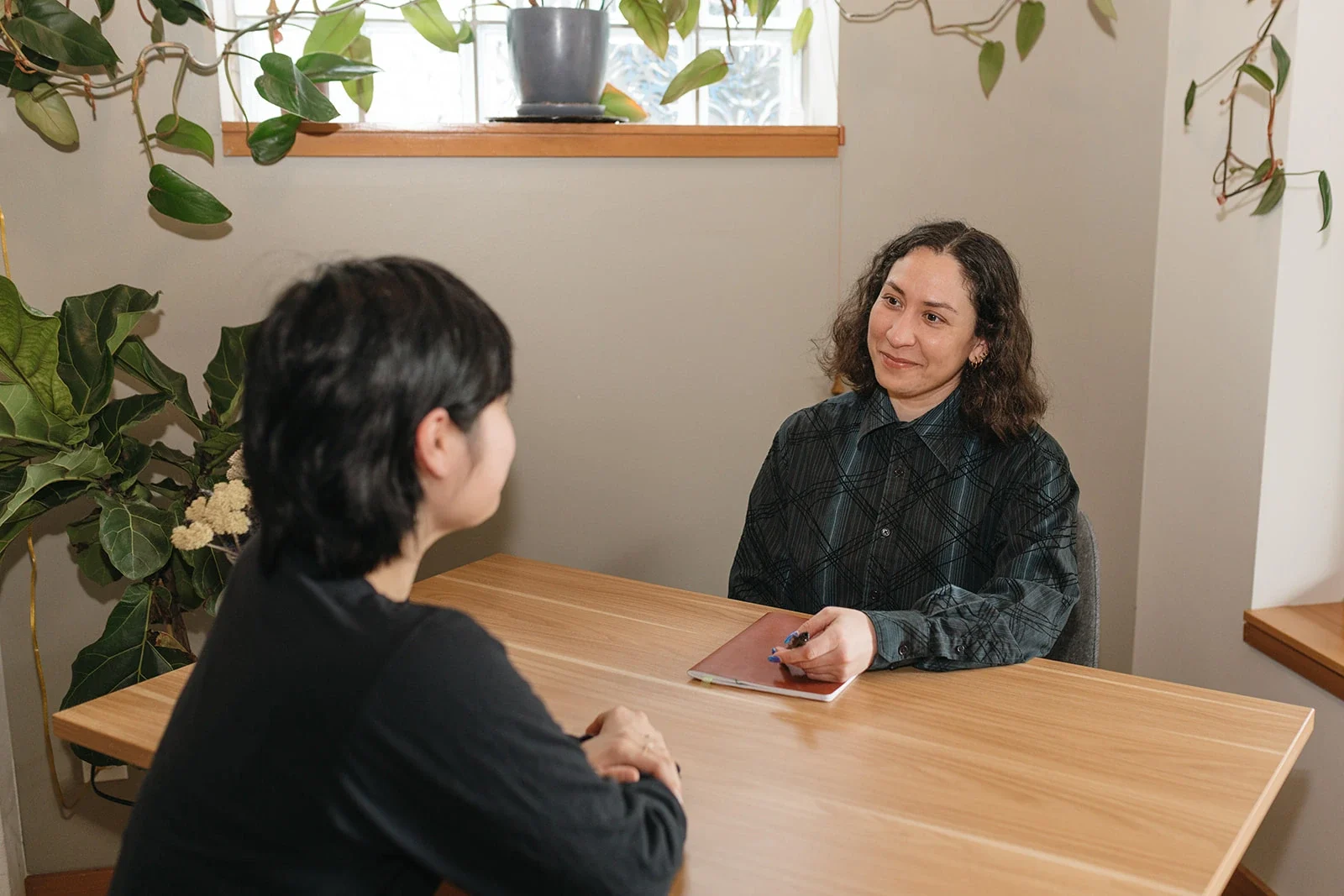 A practitioner and a client talk across a wooden table with a notebook open, in a bright room with trailing plants